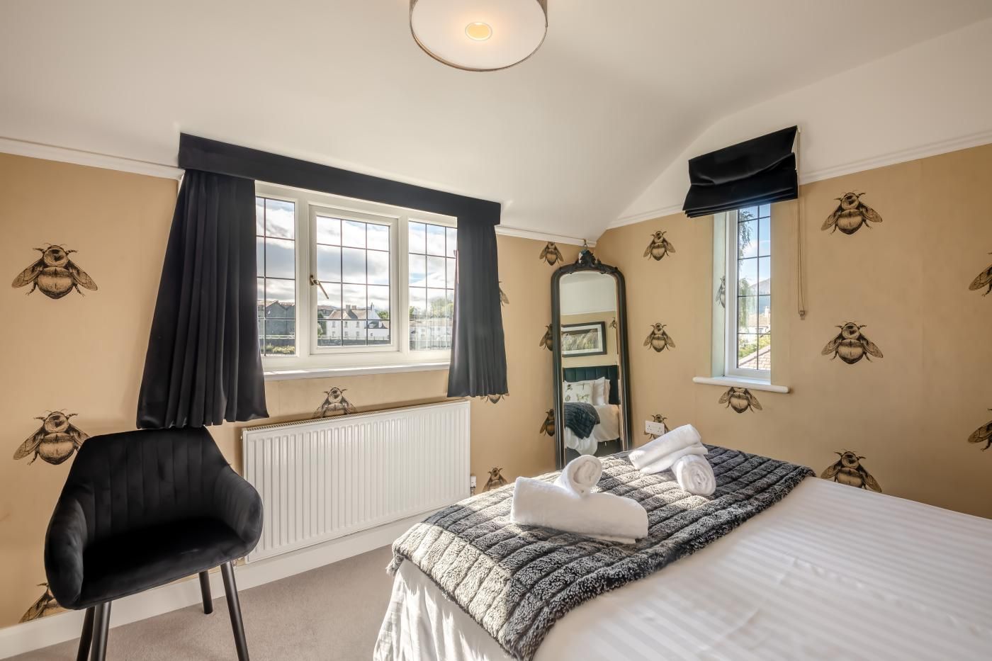 Une chambre à coucher avec un lit, une chaise, une fenêtre avec des rideaux, un radiateur et un mur recouvert d’un papier peint à motifs d’abeilles.
