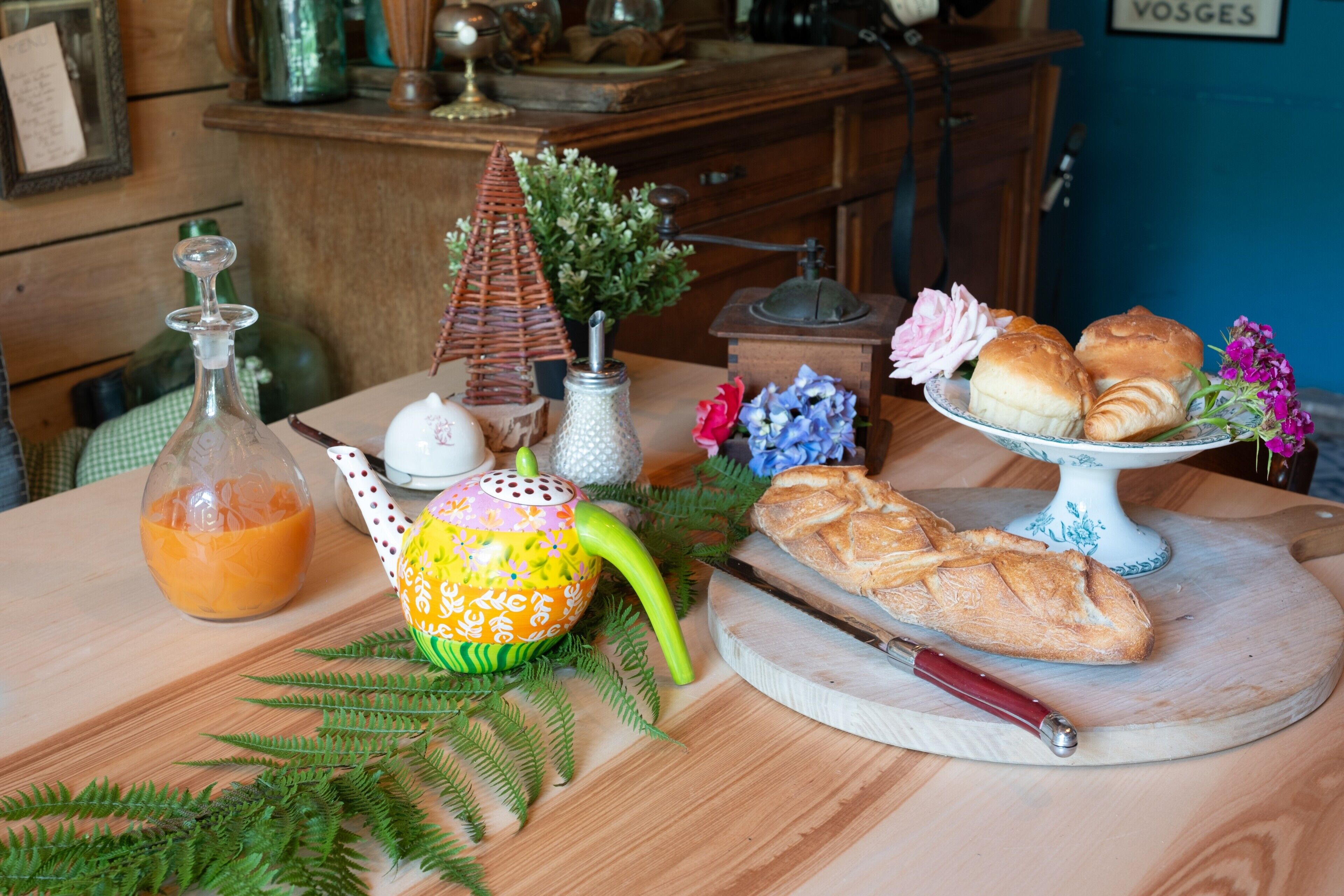 Une table en bois sur laquelle se trouvent une bouteille de jus d’orange, une théière, un petit objet en forme de pyramide, une miche de pain et une assiette de pâtisseries.