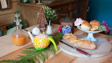 Une table en bois sur laquelle se trouvent une bouteille de jus d’orange, une théière, un petit objet en forme de pyramide, une miche de pain et une assiette de pâtisseries.