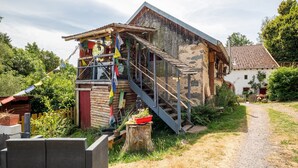 Ein rustikales Holzgebäude mit einer Treppe, die zu einem überdachten Bereich führt, umgeben von Grünpflanzen und einem Kiesweg.