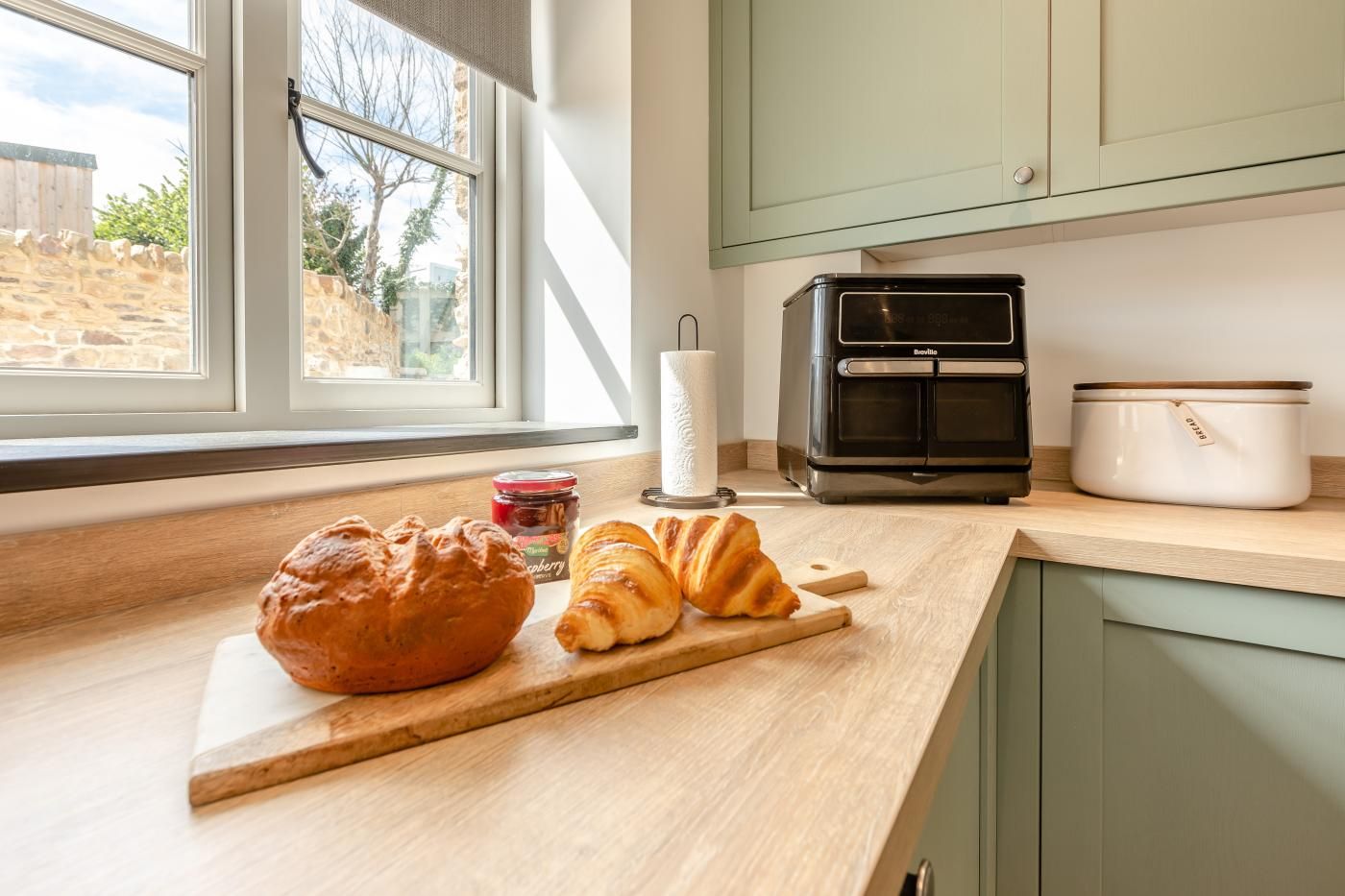 A kitchen with a loaf of bread, croissants, a jar of jam, a toaster, and a slow cooker on the counter.