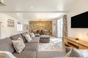 A modern living room with a stone wall, a grey sofa, a wooden TV stand, and a dining area in the background. - 3 Bed in Croyde (oc-t44822) (Croyde)