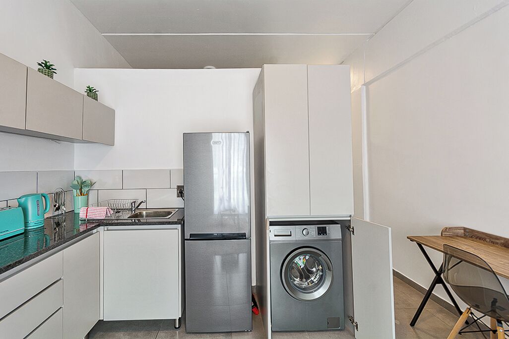 A compact kitchen with a refrigerator, washing machine, and a small dining table.