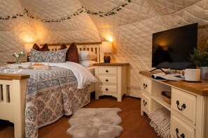 Ein gemütliches Schlafzimmer mit einer gepolsterten Decke, einem Bett mit gemusterten Betttextilien, einem Nachttisch mit Lampe, einem Fernseher und einem Schreibtisch mit einer Tasse.