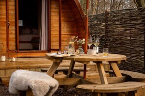 A wooden deck with a round table set for dining, a fur-covered bench, and a rustic wooden fence.