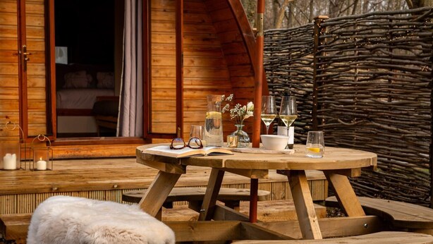 Een houten terras met een ronde eettafel, een met bont beklede bank en een rustieke houten omheining.