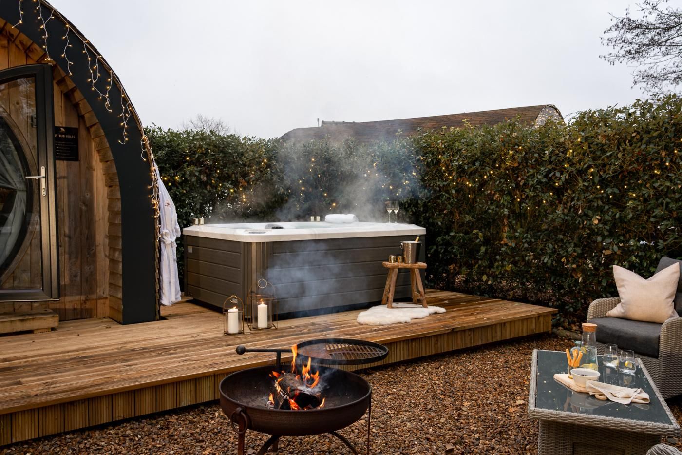 Ein Whirlpool auf einer Holzterrasse mit einem Feuerkorb, umgeben von Grünpflanzen und einer gemütlichen Sitzecke.