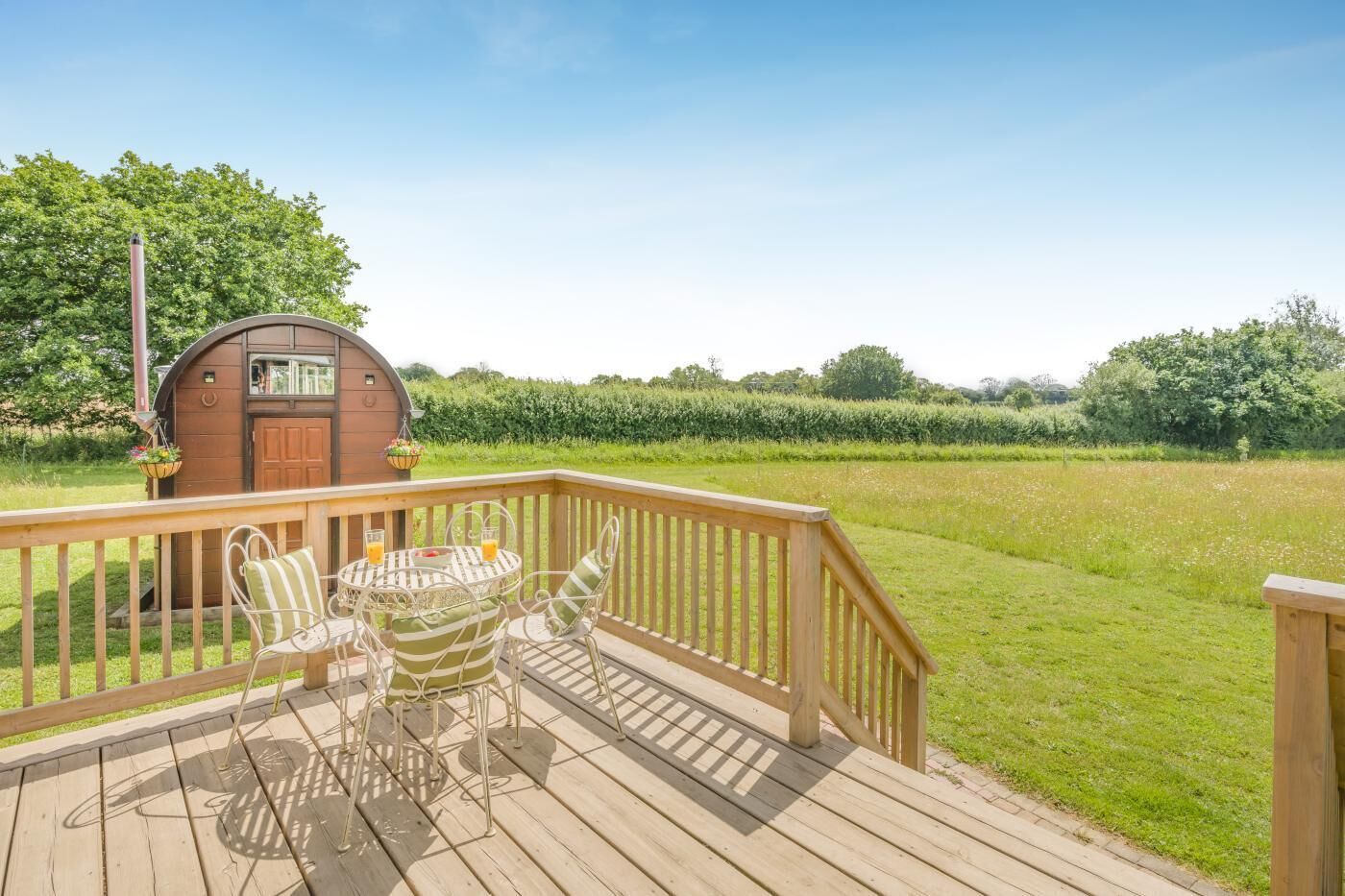 A wooden deck with a table and chairs, a small building with a door and windows, and a grassy field in the background.