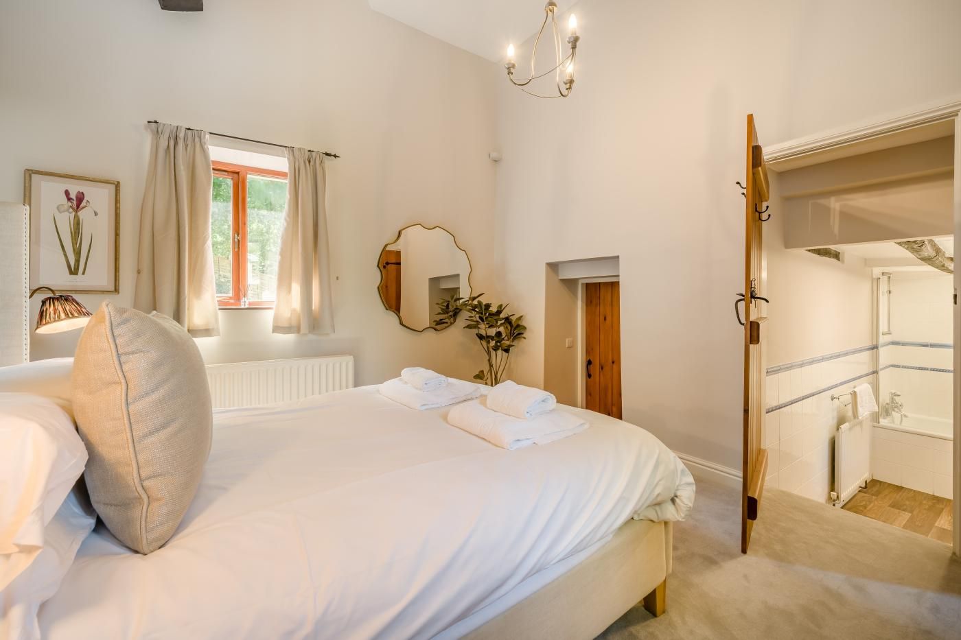 A bedroom with a large bed, a mirror, a window with curtains, and a bathroom visible through an open door.