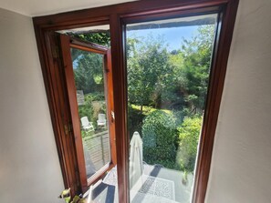 Eine Glastür führt in einen Garten mit einer Holzterrasse und weißen Stühlen.