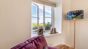 Una habitació amb una finestra gran, una làmpada de peu, un coixí morat i una taula petita amb un ram de flors.