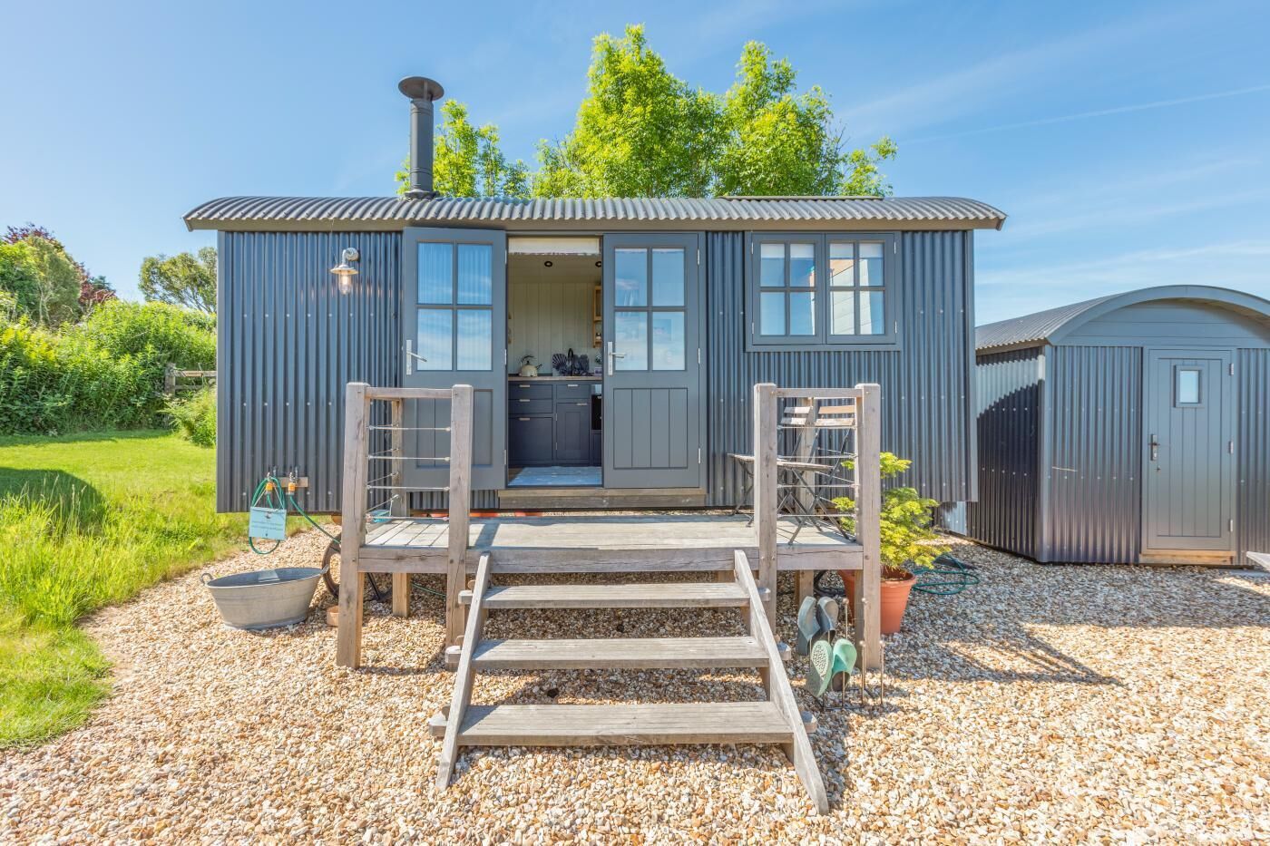 A compact, single-story cabin with a metal exterior, a wooden deck, and a gravel driveway.