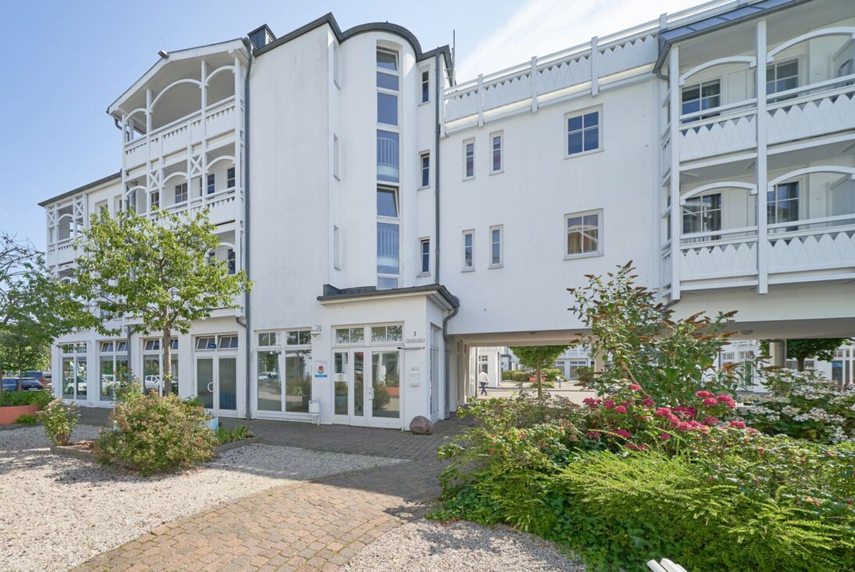 En hvid bygning med flere etager, altaner, en lukket indgang og en grusvej.