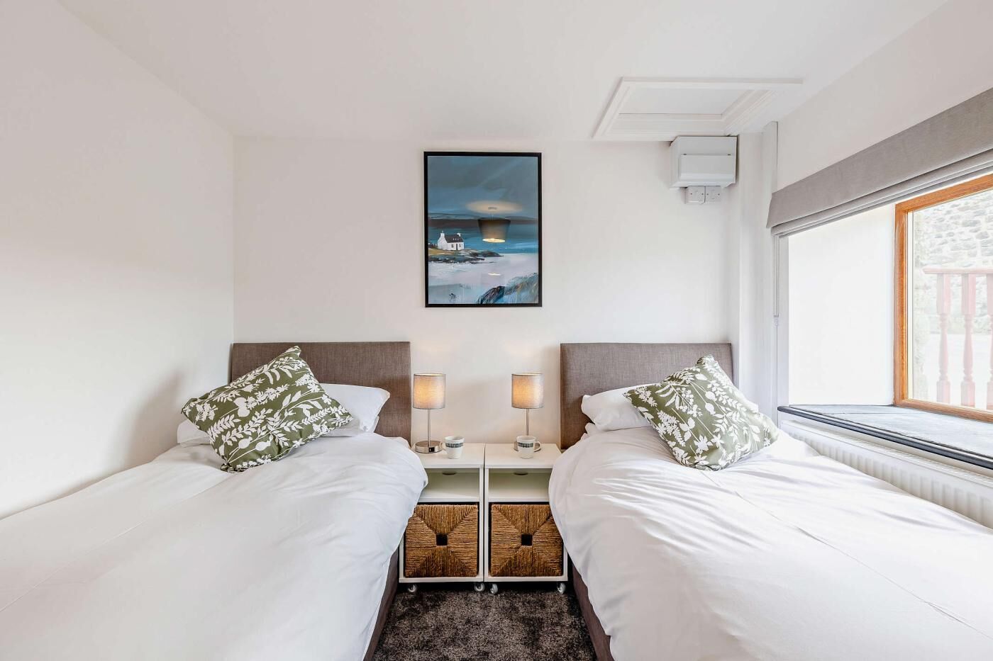 A hotel room with two beds, a nightstand with a lamp, a picture on the wall, and a window with a view.
