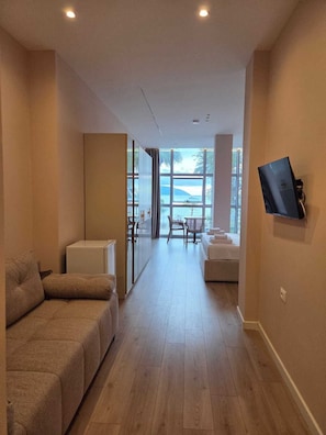 A hotel room with a sofa, a small table, a TV mounted on the wall, and a view of the sea through large windows. - Sunset Boulevard Rooms (Vlore)