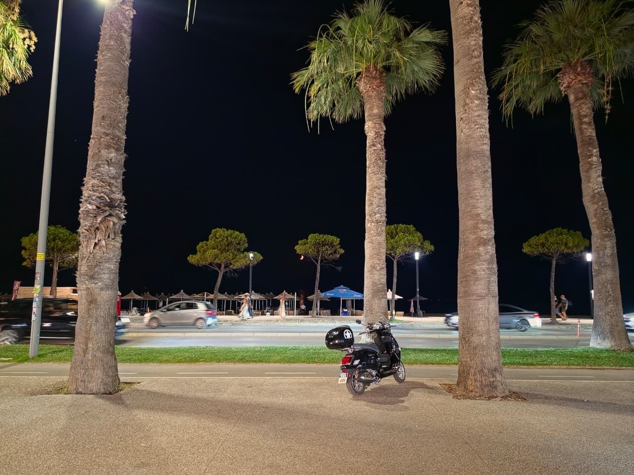 Escena nocturna con palmeras, una moto estacionada y una calle con vehículos en movimiento.