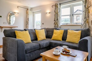 Ein modernes Wohnzimmer mit einem Ecksofa, einem Holztisch und Blick auf ein Steinhaus durch die Fenster.