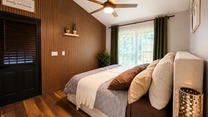 Une chambre avec un lit, un ventilateur de plafond, une fenêtre avec des rideaux, une porte et une étagère fixée au mur, ornée de plantes.
