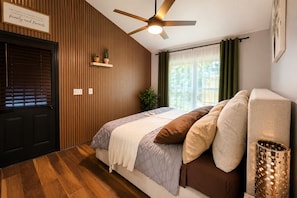 Ein Schlafzimmer mit einem Bett, einem Deckenventilator, einem Fenster mit Vorhängen, einer Tür und einem an der Wand befestigten Regal mit Pflanzen.