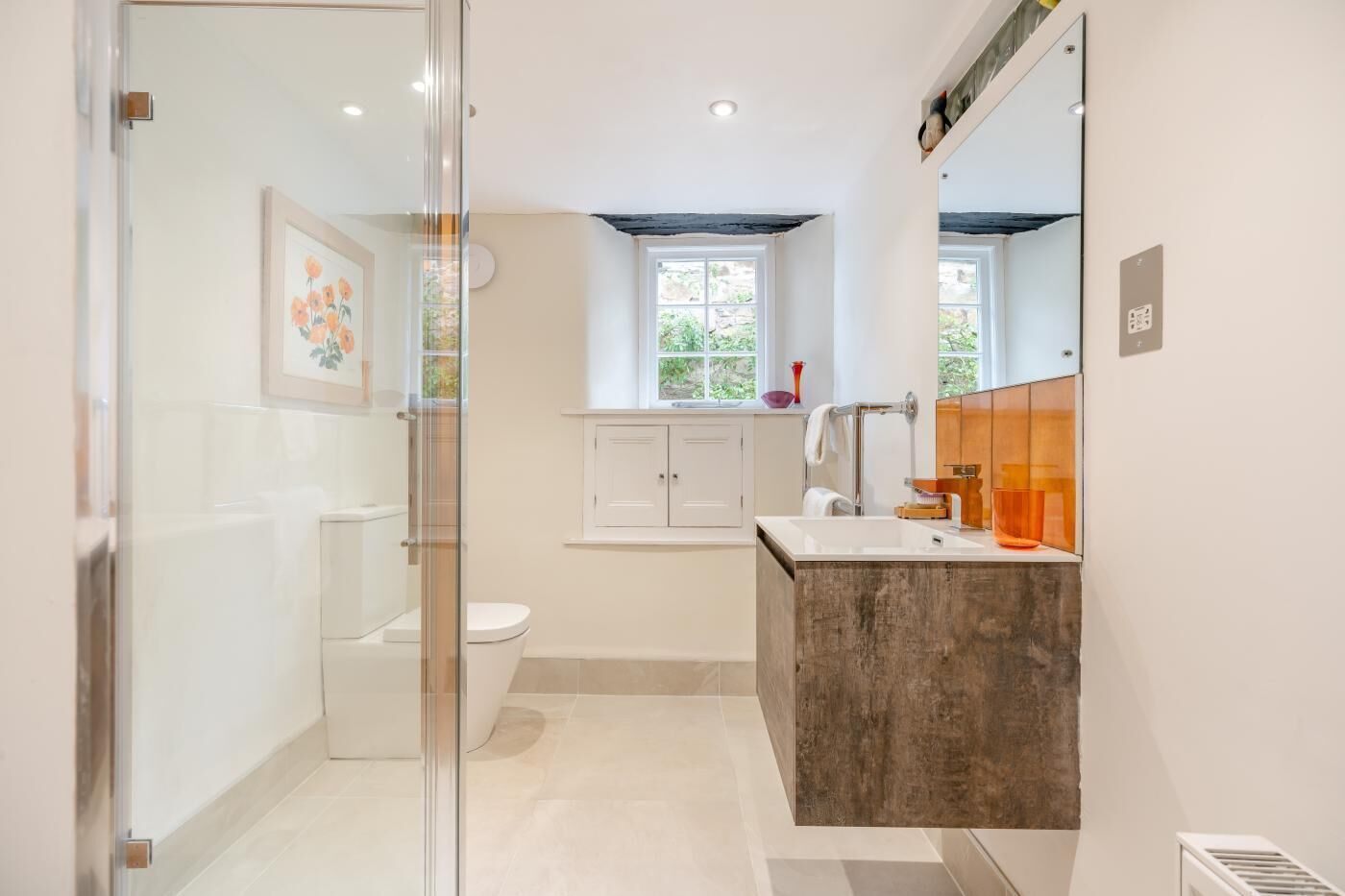 Un bagno moderno con box doccia in vetro, un water, un lavandino con mobile contenitore in legno e una finestra che offre luce naturale.
