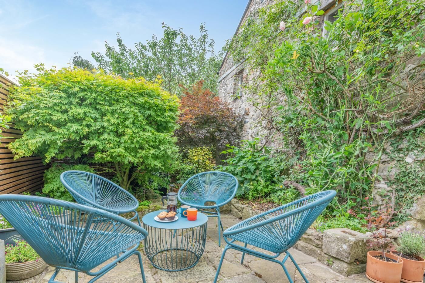 A patio with blue chairs and a table, surrounded by greenery and a stone wall.