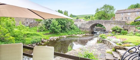 Vista al río con un puente de piedra, abundante vegetación y un área de patio con sillas y una sombrilla.