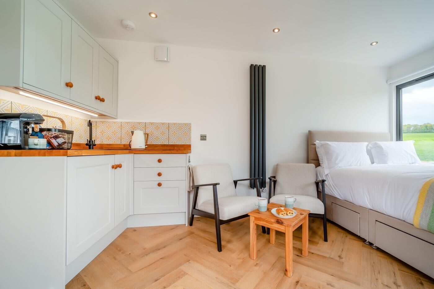 A compact room with a kitchenette, two chairs, a small table with a plate and mug, a bed with white bedding, and a window with a view.