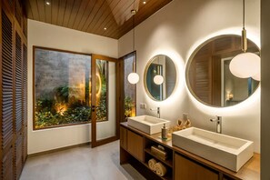 Une salle de bain moderne avec deux lavabos, un grand miroir et une vue sur un jardin, visible à travers une porte en verre.