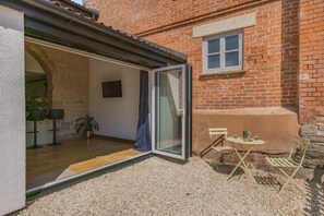Eine offene Terrasse mit Tisch und Stühlen, eine Glastür zu einem Innenraum mit Pflanze und Barhocker.