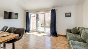 Een ruime woonkamer met houten vloer, een bank, een eettafel met stoelen en een schuifpui naar een buitenruimte.