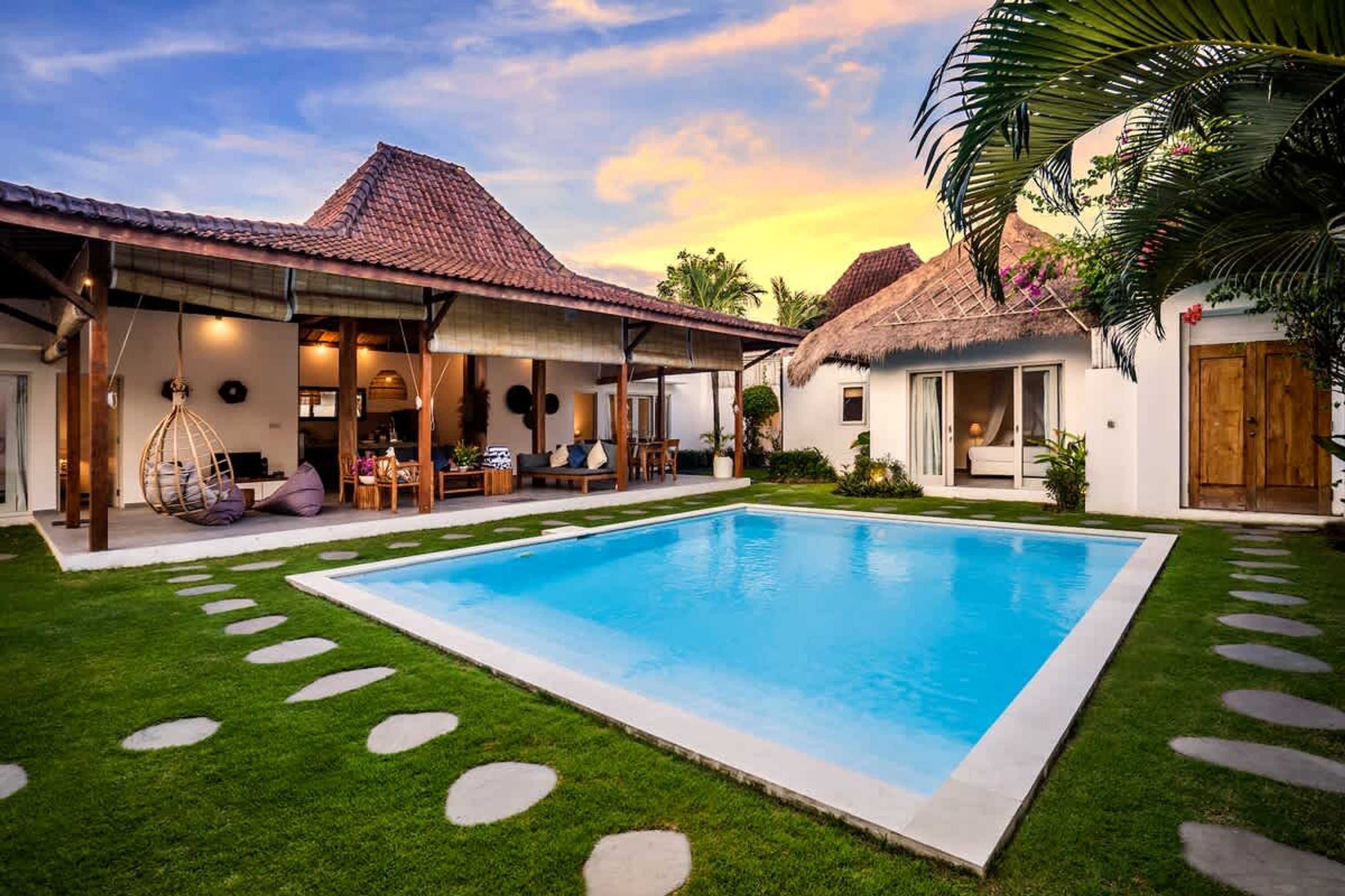 A poolside area with a clear blue pool, a covered seating area, and a thatched-roof structure.