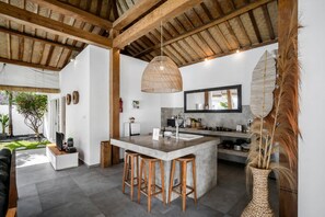 A modern kitchen with a central island, wooden stools, a hanging light fixture, and a large mirror above the sink. - Villa Nakal in Seminyak (Seminyak)