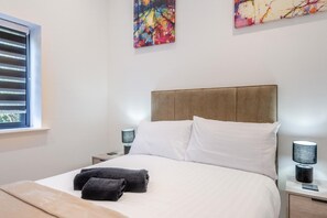 Una cama bien hecha con sábanas blancas y dos toallas negras, un cabecero de madera, lámparas de noche y arte abstracto en las paredes.