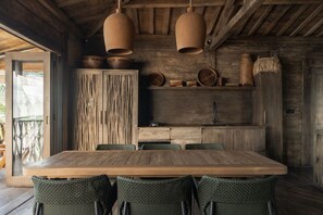 Um interior rústico em madeira com uma grande mesa de jantar, cadeiras de vime e uma cozinha com armários de madeira e panelas penduradas.