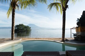 A clear view of a swimming pool with lounge chairs, palm trees, and a distant view of a body of water and a mountain. - Sunset Beachwood Villa (Nusa Lembongan)