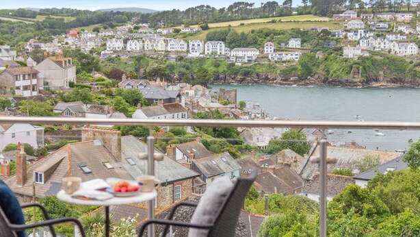 A balcony with a table and chairs overlooking a coastal town with houses and a body of water. - 4 Bed in Polruan (oc-s41411) (Fowey)