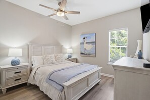 Une chambre à coucher avec un lit, des tables de chevet, un ventilateur de plafond, un tableau et une fenêtre avec des stores.