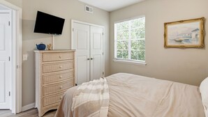 A bedroom with a bed, a television mounted on the wall, a dresser, a window with blinds, and a framed picture on the wall. - Coastal Condo: Pet friendly/Pool/Mins to UNCW, Downtown & Wrightsville Beach (Wilmington)