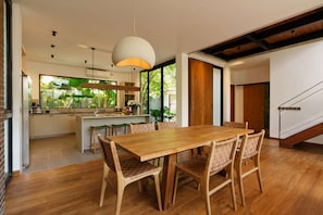 A modern kitchen with a central island, wooden dining table with woven chairs, and a large window overlooking greenery. - Villa Pakuk in Kecamatan Kuta Utara (Kecamatan Kuta Utara)