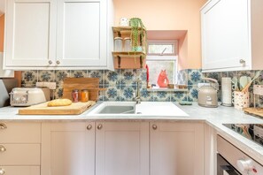 A kitchen with white cabinets, a wooden countertop, a toaster, a bread loaf, jars, a sink, and a window with a plant. - 2 Bed in Great Broughton (oc-m44944) (Great Broughton)