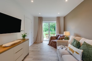 A modern living room with a sofa, a coffee table, a TV mounted on the wall, and a dining area visible through a glass door.
