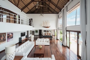 A spacious living area with a high ceiling, a sailboat decoration, a white sofa, a coffee table, and a TV. - Villa Blanca in Manggis (Manggis)