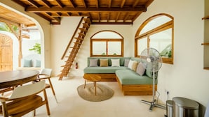 Ein gemütliches Wohnzimmer mit Holzbalkendecke, einem grünen Sofa mit Kissen, einem runden Holztisch und einem Ventilator.