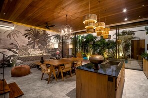 Ein modernes Interieur mit Holzbalkendecke, einem Essbereich mit Holztisch und -stühlen, einer Kücheninsel und einem Wandbild von tropischen Pflanzen.