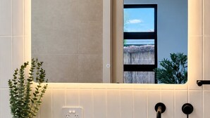 Ein Badezimmer mit einem großen Spiegel, einer Topfpflanze und Blick auf ein reetgedecktes Gebäude durch das Fenster.