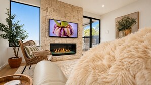 Ein Wohnzimmer mit Kamin aus Stein, einem an der Wand montierten Fernseher, Korbmöbeln und einer großen Kübelpflanze.