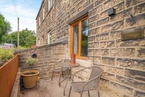A stone building with a small patio area featuring a table and chairs, a potted plant, and a wooden planter. - 2 Bed in Holmbridge (oc-s44882) (Holmfirth)