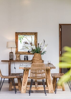 A rustic wooden dining table with chairs, a woven vase with flowers, a framed mirror, and a lamp on a shelf. - Villa Namu in Seminyak (Seminyak)