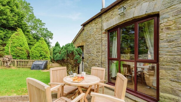 Een stenen gebouw met een groot raam, een ronde houten tafel met stoelen en een barbecue in de tuin.