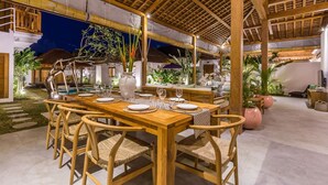 Un área de comedor al aire libre con mesas y sillas de madera, rodeada de plantas y una piscina.